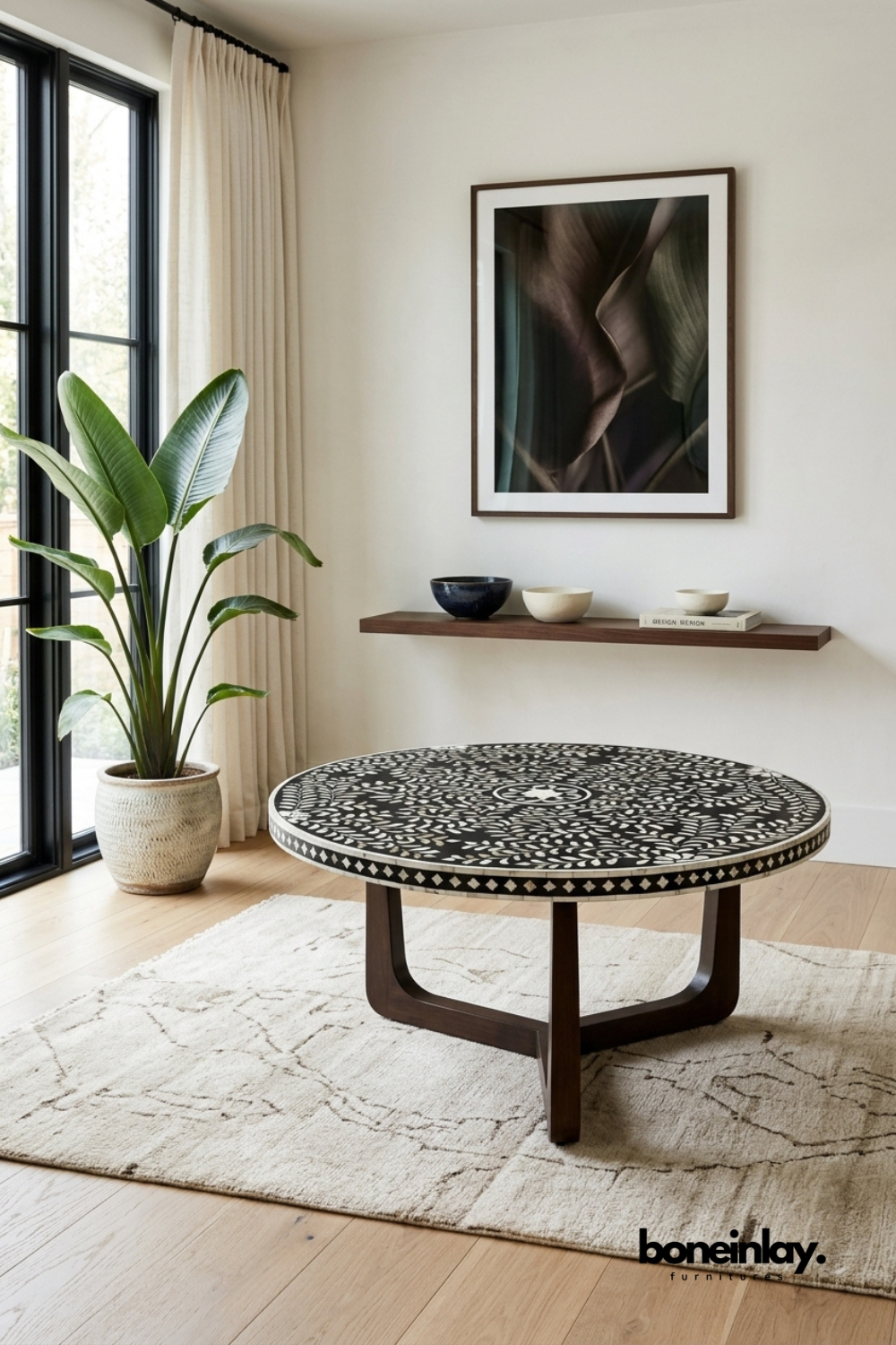 Round black and white bone inlay coffee table featuring an intricate floral vine pattern with a mosaic tile border edge. Sculptural walnut wood base with curved legs adds warmth and structural support. Styled with a potted bird of paradise plant beneath a floating wood shelf with ceramic bowls. A striking round coffee table for modern and eclectic living spaces.