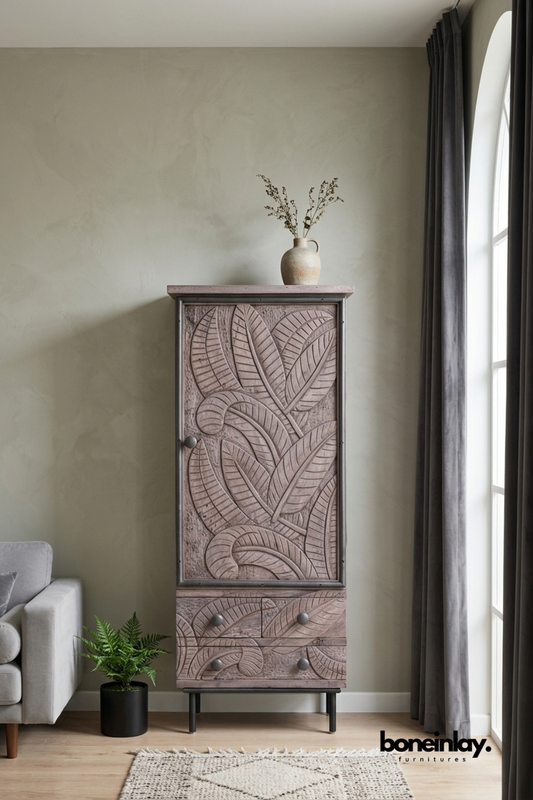 Tall wood cabinet with a hand-carved tropical leaf pattern door.
Two carved drawers sit below the single door for extra storage.
Framed in a dark metal edge with round metal legs and pulls.
Styled beside a fern plant and grey linen sofa in a modern living room.