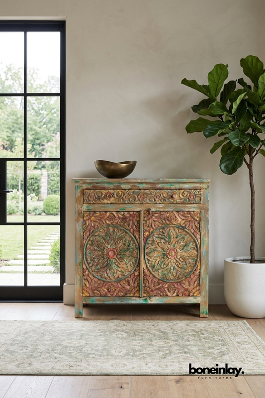 Natural mango wood accent cabinet featuring intricate floral and botanical hand-carved detailing across doors and drawer front. Includes a single top drawer with ring pulls and double carved doors with traditional flower urn motifs. Styled with a ceramic vase, dried botanicals, and stacked design books beneath framed abstract wall art. A statement storage piece bringing artisanal warmth to entryways and living rooms.