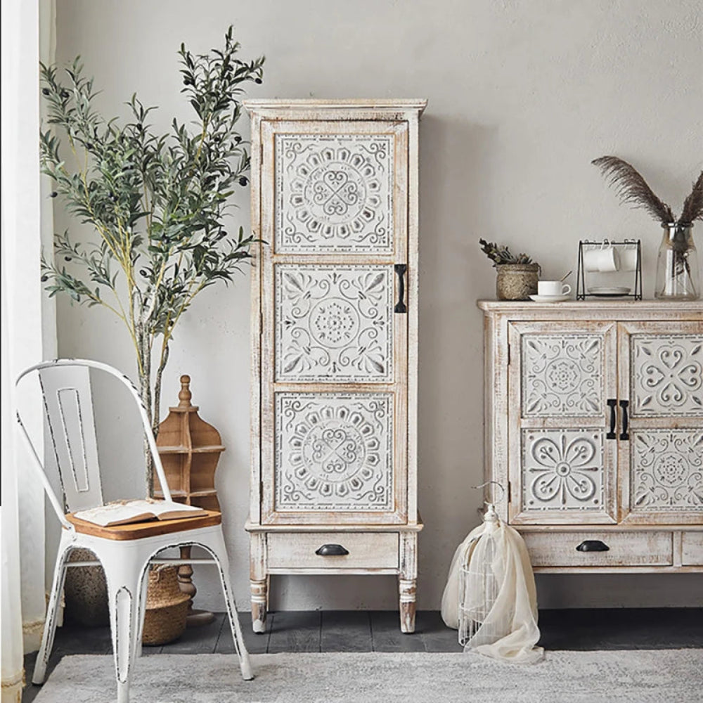 White floral accent cabinet with a tall carved door and bottom drawer, styled in a bright room beside a chair.