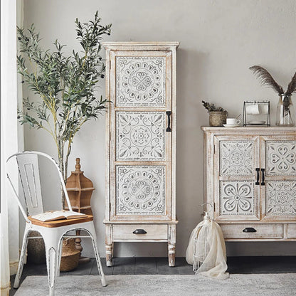 White floral accent cabinet with a tall carved door and bottom drawer, styled in a bright room beside a chair.