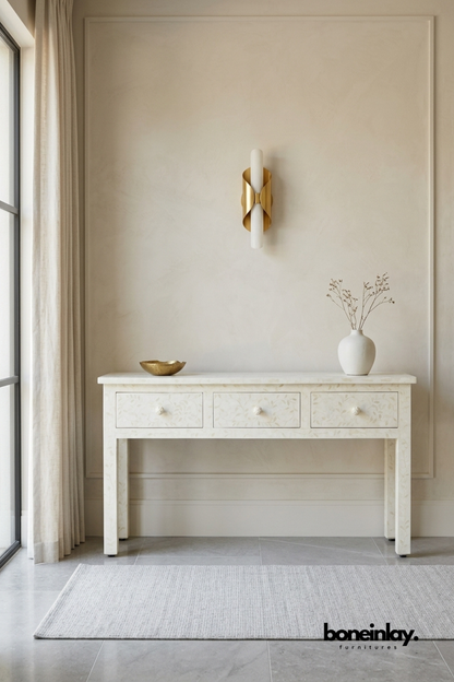 Ivory bone inlay console table featuring a delicate all-over floral vine pattern across the top and three drawers. Textured bone knobs and clean tapered legs complete the refined, tailored silhouette. Styled with a brass dish and ceramic vase beneath a sculptural brass wall sconce. An elegant console table for entryways and hallway styling.