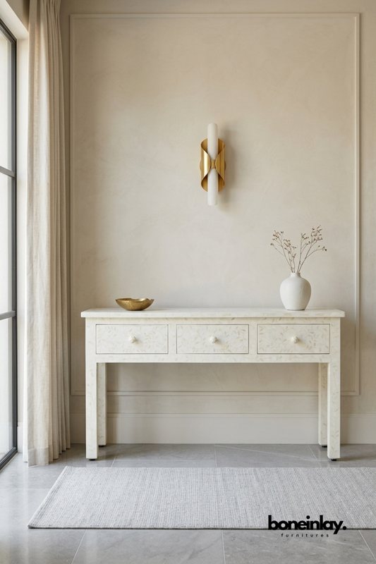 Ivory bone inlay console table featuring a delicate all-over floral vine pattern across the top and three drawers. Textured bone knobs and clean tapered legs complete the refined, tailored silhouette. Styled with a brass dish and ceramic vase beneath a sculptural brass wall sconce. An elegant console table for entryways and hallway styling.