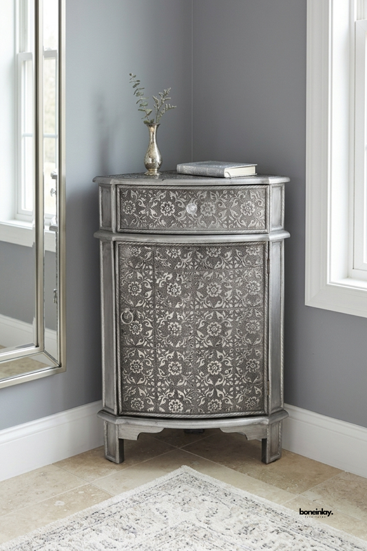 Handcrafted silver metal corner accent cabinet featuring intricate hand-embossed floral tile patterns across the single curved drawer and lower cupboard door. Styled nestled in a cozy room corner beside a mirror and window, this vintage-inspired metallic nightstand maximizes small spaces while adding rustic luxury and ornate bohemian charm.