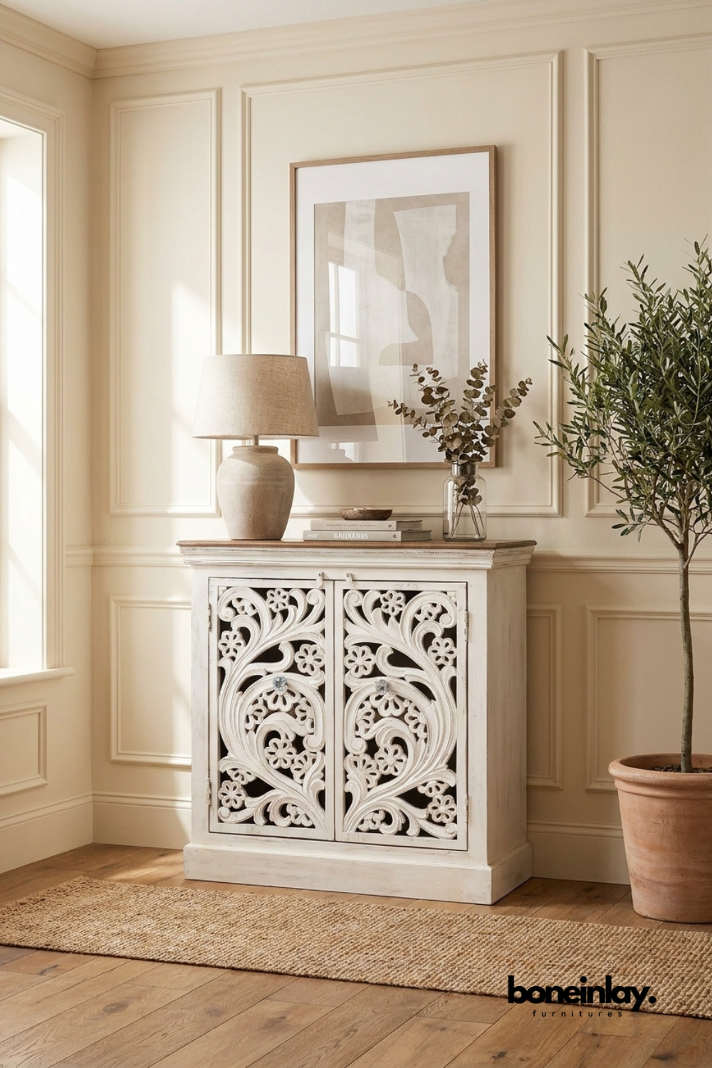White carved wood accent cabinet with a flowing scroll and floral pattern.
Two doors feature open lattice carving with glass-bead style knobs.
Natural wood tabletop tops the distressed white cabinet base.
Styled beneath a framed print with a ceramic lamp and eucalyptus stems.