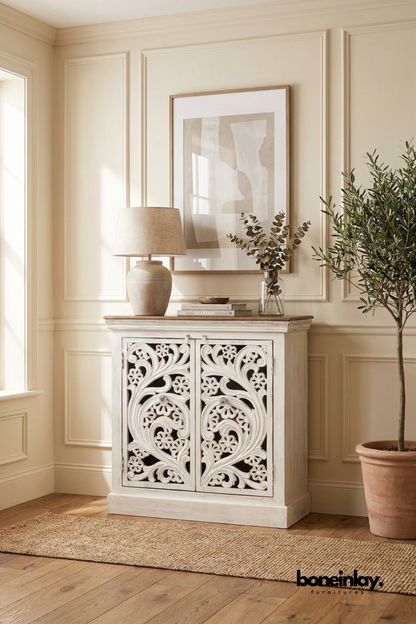 White carved wood accent cabinet with a flowing scroll and floral pattern.
Two doors feature open lattice carving with glass-bead style knobs.
Natural wood tabletop tops the distressed white cabinet base.
Styled beneath a framed print with a ceramic lamp and eucalyptus stems.