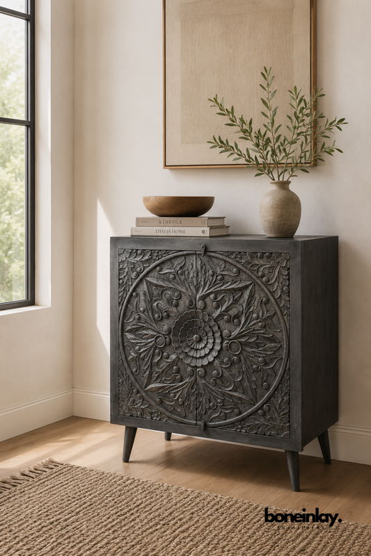 Grey carved wood cabinet with a large round floral medallion design.
Detailed scroll and petal carving covers the full cabinet door front.
Tapered wood legs give it a light mid-century silhouette.
Styled with a wood bowl and stacked books on a jute rug.