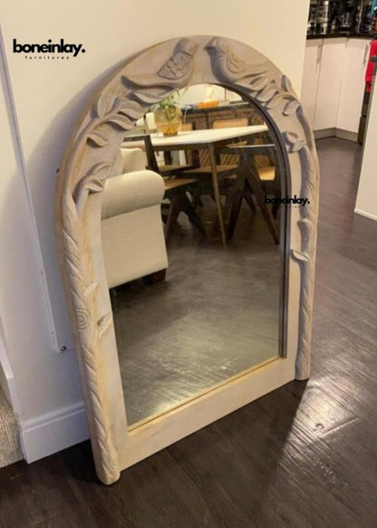 Wooden ornithology mirror with a carved bird frame, shown at an angle beside a table in a furnished room.