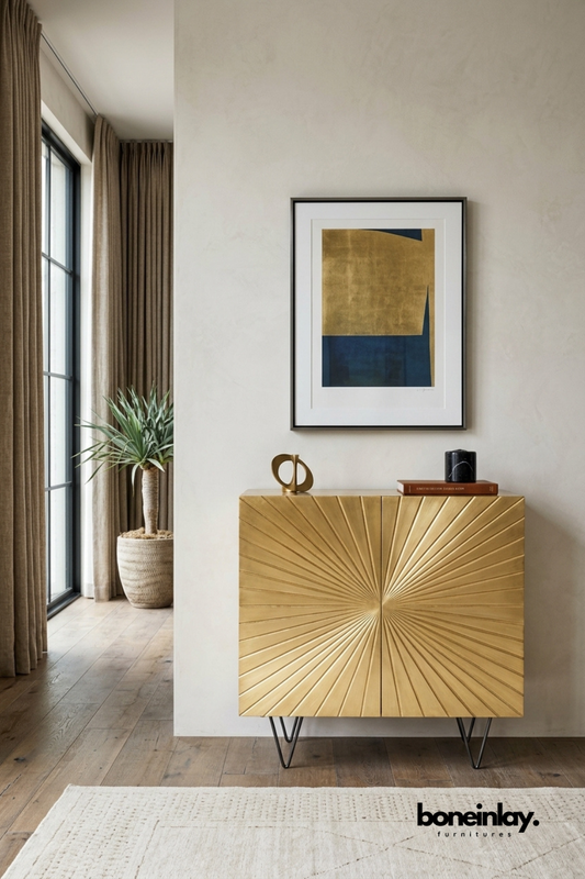 Brass gold sunburst accent cabinet featuring a radiating ribbed pattern carved across double doors. Elevated on sleek black hairpin legs for a glamorous mid-century modern touch. Styled with a brass sculpture and leather book beneath framed abstract wall art. A luxe statement cabinet for adding warmth and shine to entryways or living rooms.