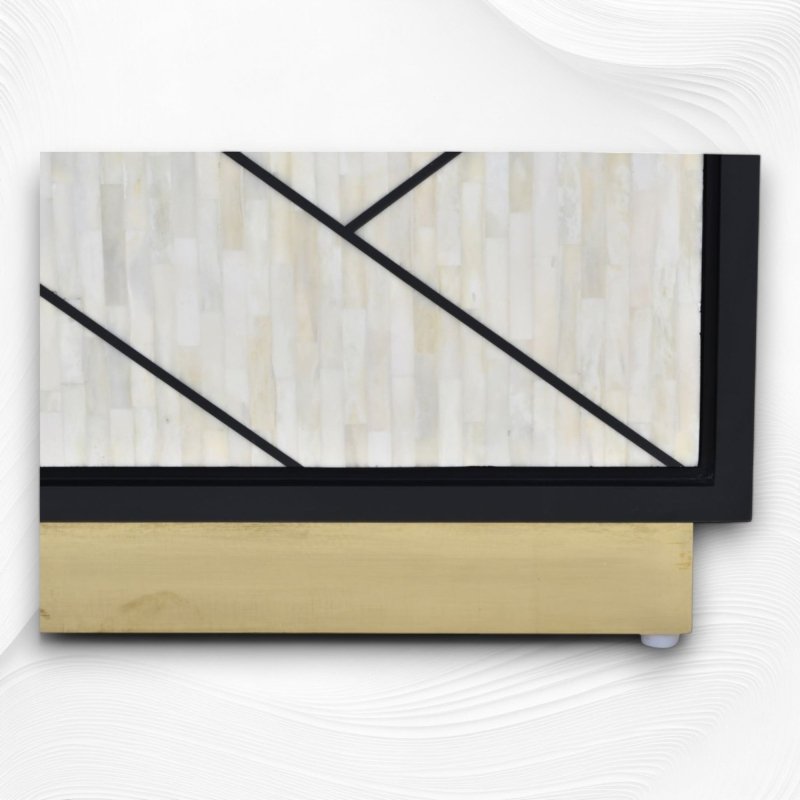 Macro detail of the polished brass gold base plinth of a bone inlay credenza cabinet with ivory geometric panel inlay and black mango wood frame junction visible.