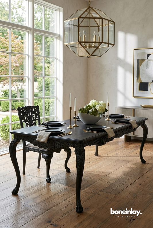A sophisticated dining room featuring a black menagerie hand-carved wooden dining table with ornate cabriole legs, styled with gold candles and white flowers under a large geometric gold pendant light.