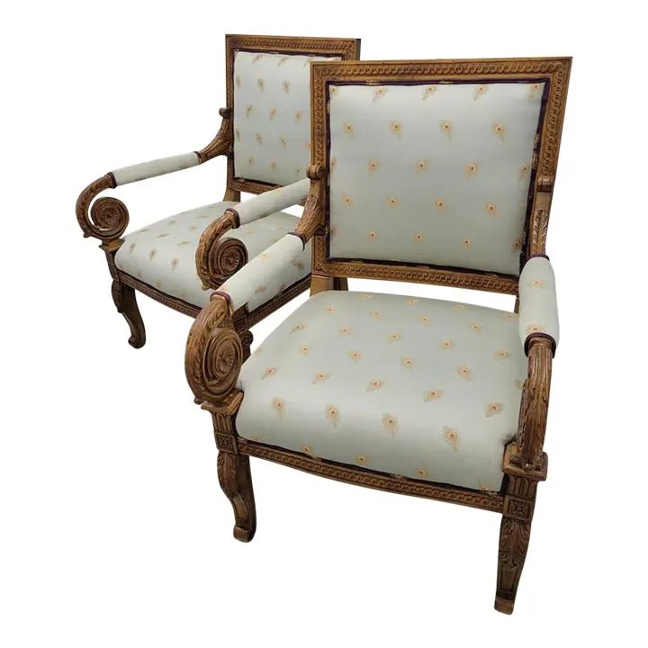 A pair of ornate, hand-carved solid wood Empire style bergère chairs with ivory patterned upholstery and intricate scrolling details.