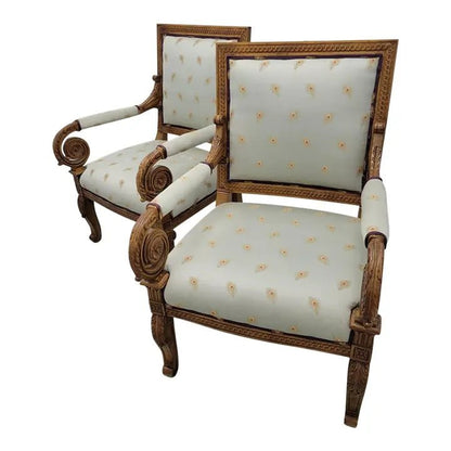 A pair of ornate, hand-carved solid wood Empire style bergère chairs with ivory patterned upholstery and intricate scrolling details.