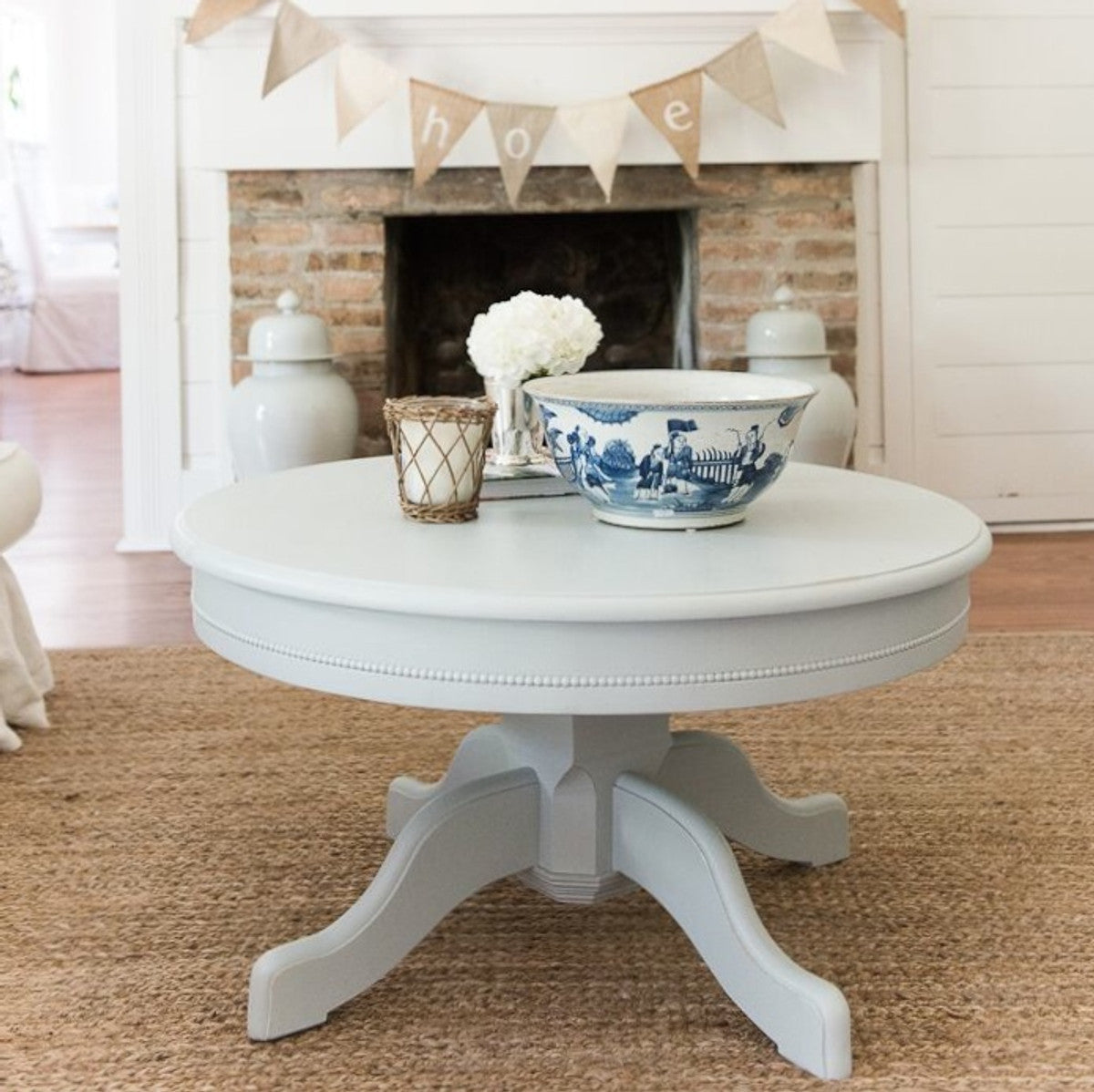  French-style round pedestal coffee table with beaded trim, styled with a blue-and-white bowl, flowers and a candle.