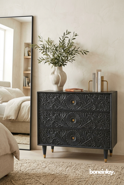 Gulliver three-drawer credenza in a charcoal finish, featuring intricate floral and bird hand-carvings, gold-toned hardware, and tapered legs with gold-dipped feet in a modern bedroom.