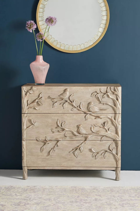 A 3-drawer solid wood dresser with intricate hand-carved reliefs of birds and tree branches, finished in a light natural wash with tree-trunk style legs.