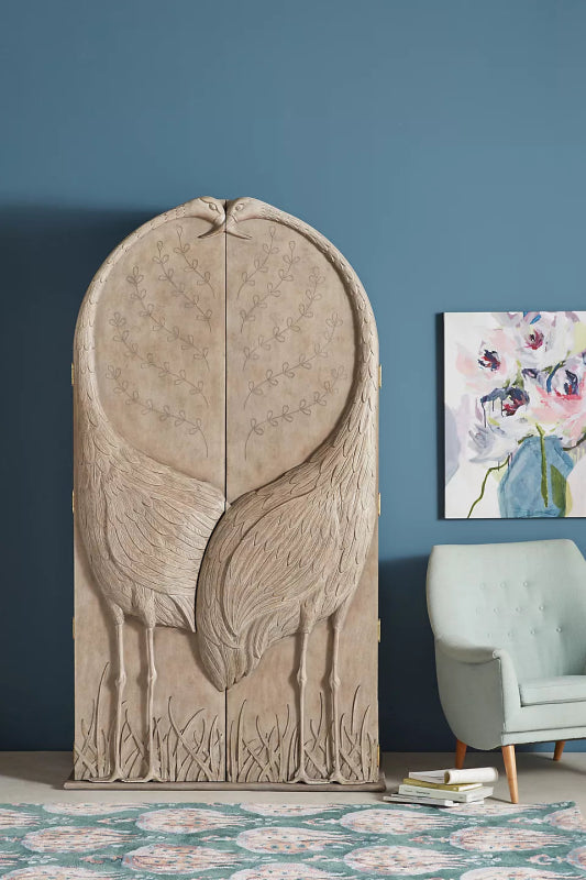 Hand carved wooden armoire with intricate crane design in a natural weathered finish against a blue wall.