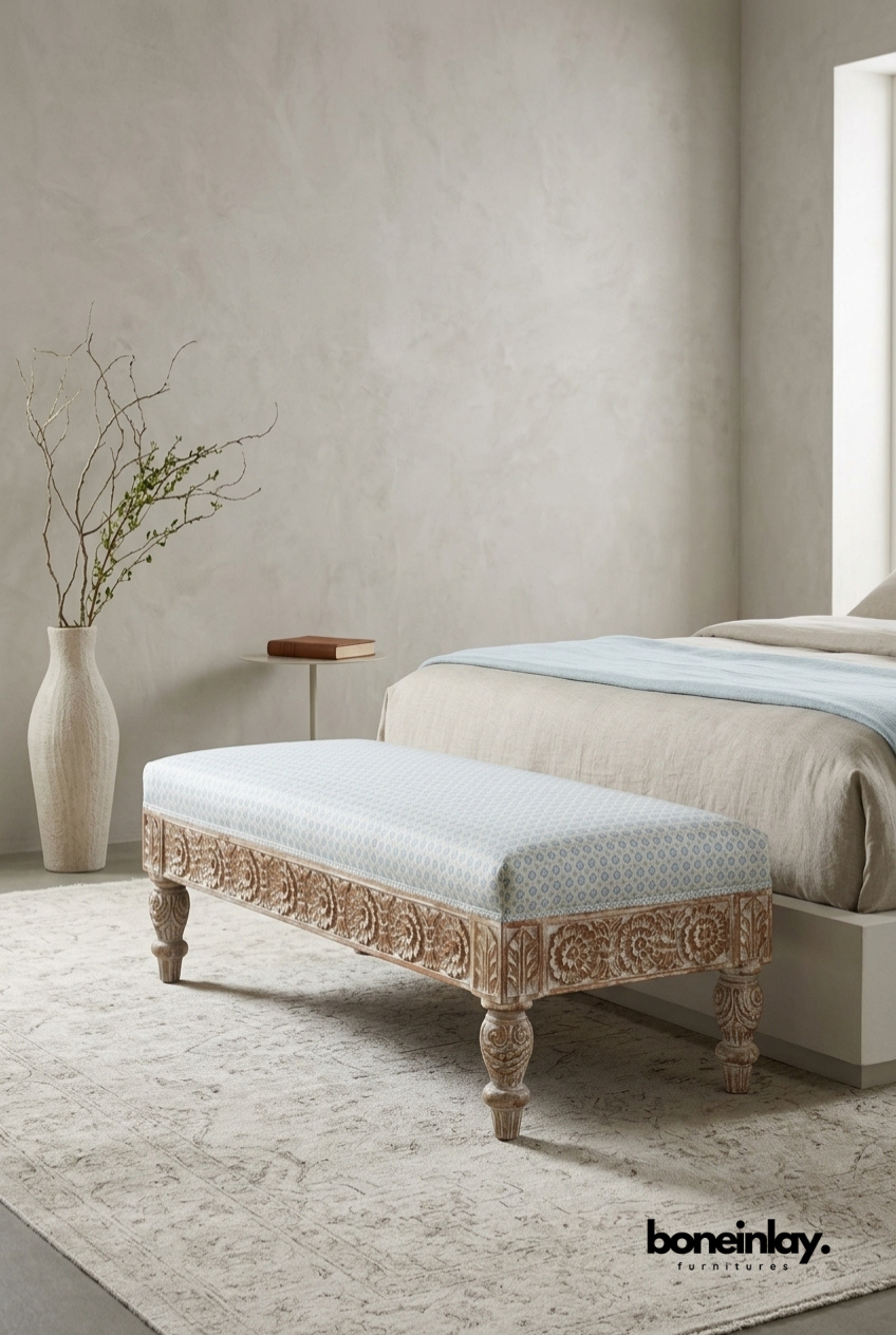 An artisanal hand-carved solid wood bench with detailed floral relief on the base and turned legs, topped with a soft, patterned upholstered cushion at the foot of a bed.