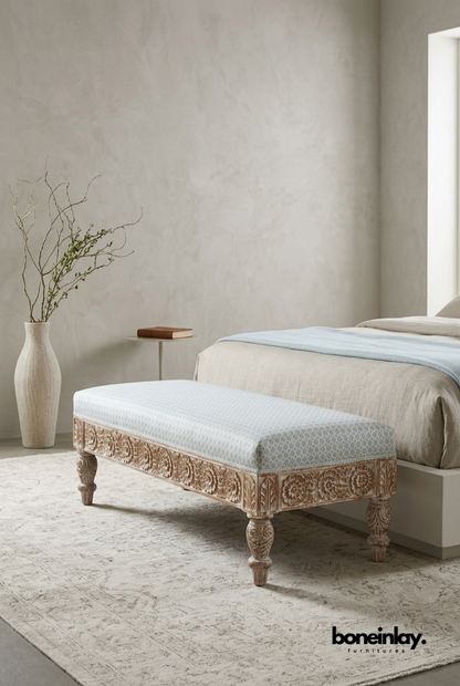 An artisanal hand-carved solid wood bench with detailed floral relief on the base and turned legs, topped with a soft, patterned upholstered cushion at the foot of a bed.