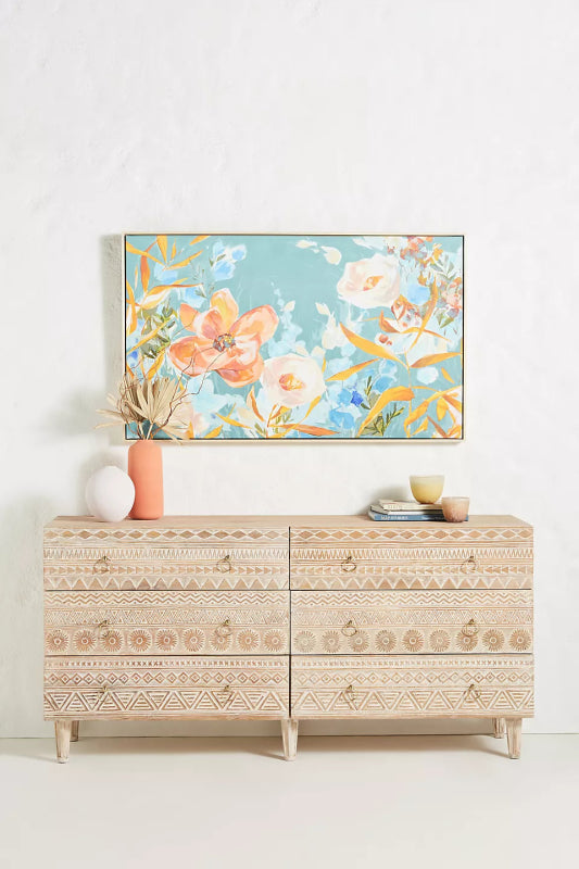 A 6-drawer solid wood dresser with hand-carved tribal geometric patterns and brass ring pulls, styled with a floral painting and modern decor in a bright room.