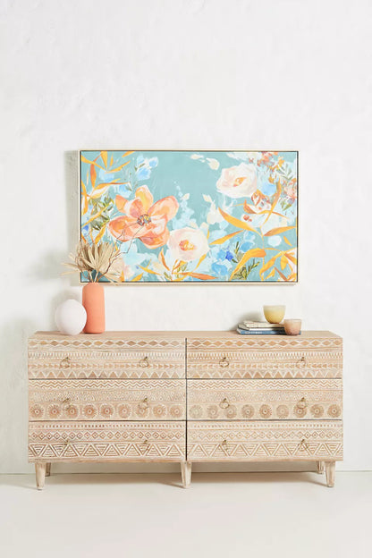 A 6-drawer solid wood dresser with hand-carved tribal geometric patterns and brass ring pulls, styled with a floral painting and modern decor in a bright room.