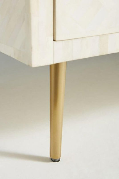 Close-up of a single tapered gold brass leg on a white optical bone inlay chest of drawers, highlighting the quality metal finish and solid wood base construction.