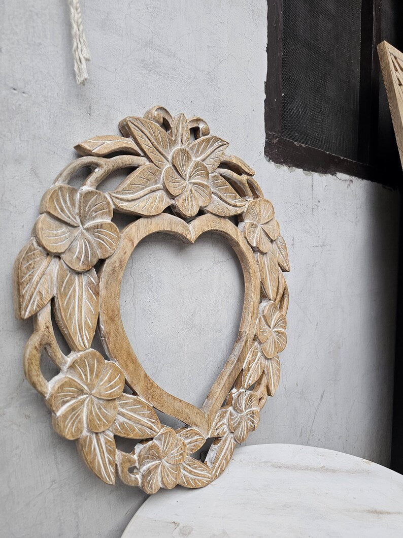 Intricate hand-carved solid wood heart-shaped mirror frame featuring blooming hibiscus flowers and a rustic natural finish against a light background.