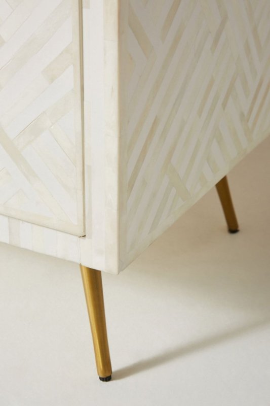 Corner detail of a white optical bone inlay 9-drawer dresser showing two slim gold tapered brass legs and the geometric bone inlay pattern on the drawer fronts and side panel.