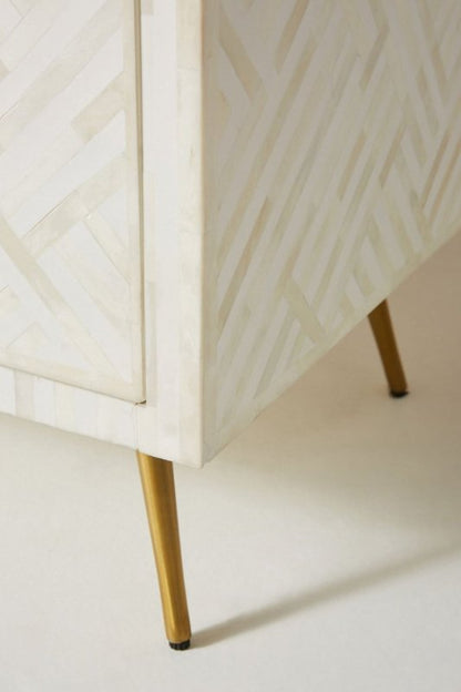 Corner detail of a white optical bone inlay 9-drawer dresser showing two slim gold tapered brass legs and the geometric bone inlay pattern on the drawer fronts and side panel.