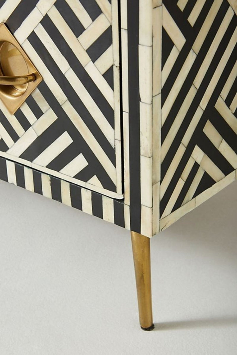 Corner close-up of a black and white optical bone inlay chest of drawers highlighting the diamond brass pull hardware, geometric stripe inlay pattern, and slim gold tapered leg.