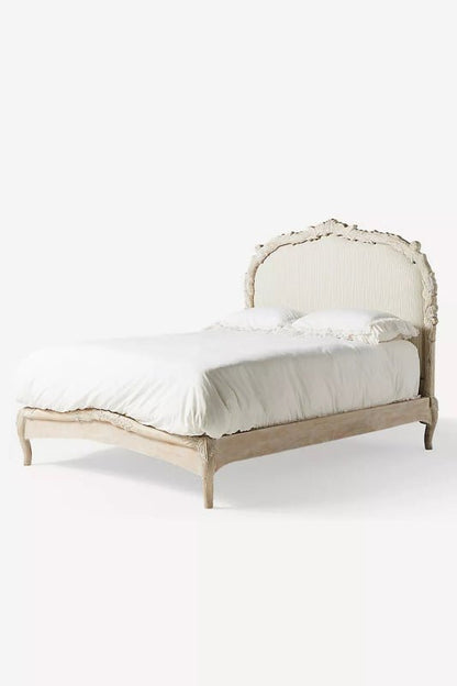 A perspective view of the Menagerie bed frame, showcasing the elegant curves of the French-inspired silhouette. The solid wood frame features a weathered natural finish with delicate floral carvings along the arched headboard, paired with clean, cream-toned upholstery for a timeless, artisanal look.
