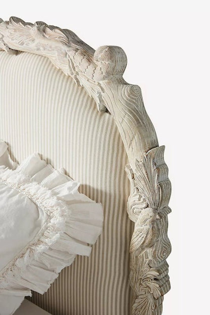 A detailed macro shot of the Menagerie headboard, focusing on the intricate hand-carved wood trim. The craftsmanship reveals ornate rose and leaf motifs surrounding the soft, piped edges of the fabric upholstery, emphasizing the high-end, handcrafted nature of the piece.