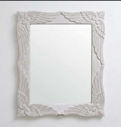 A rectangular wooden wall mirror from the Menagerie collection, featuring a carved frame with ornate wing and scrollwork patterns finished in distressed off-white and gold, shown against a plain white wall.