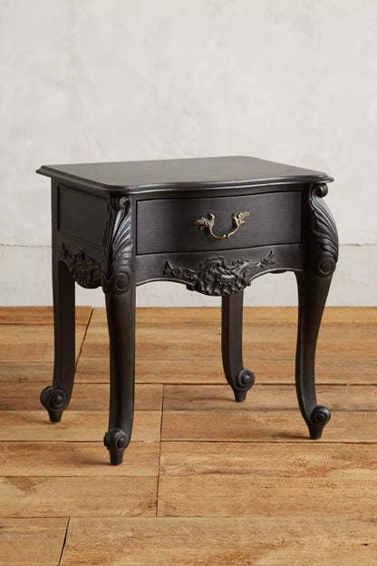  A three-quarter view of the Menara nightstand, featuring a single drawer with a vintage brass pull handle and elaborate carvings on the legs and apron.