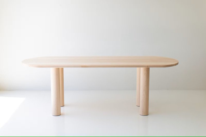 A luxury solid wood oval dining table with a natural light timber finish and thick cylindrical architectural legs.