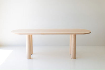 A luxury solid wood oval dining table with a natural light timber finish and thick cylindrical architectural legs.