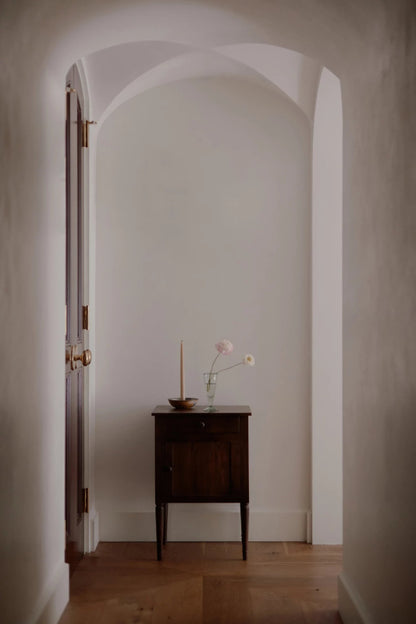  Natural finish mid-century modern cabinet table styled with a candle and flowers in a white arched hallway.