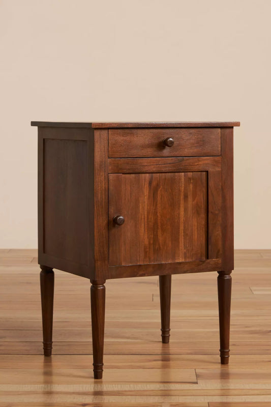 Angled view of a natural finish mid-century bedside table showing its wooden side panel, drawer, cabinet door, and tapered legs.