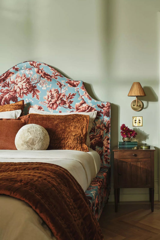 Natural finish mid-century bedside table beside a floral upholstered bed, styled with books and flowers beneath a wall lamp.