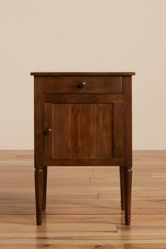 Front view of a natural finish mid-century bedside table with one drawer, a paneled cabinet door, and tapered legs.