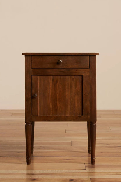 Front view of a natural finish mid-century bedside table with one drawer, a paneled cabinet door, and tapered legs.