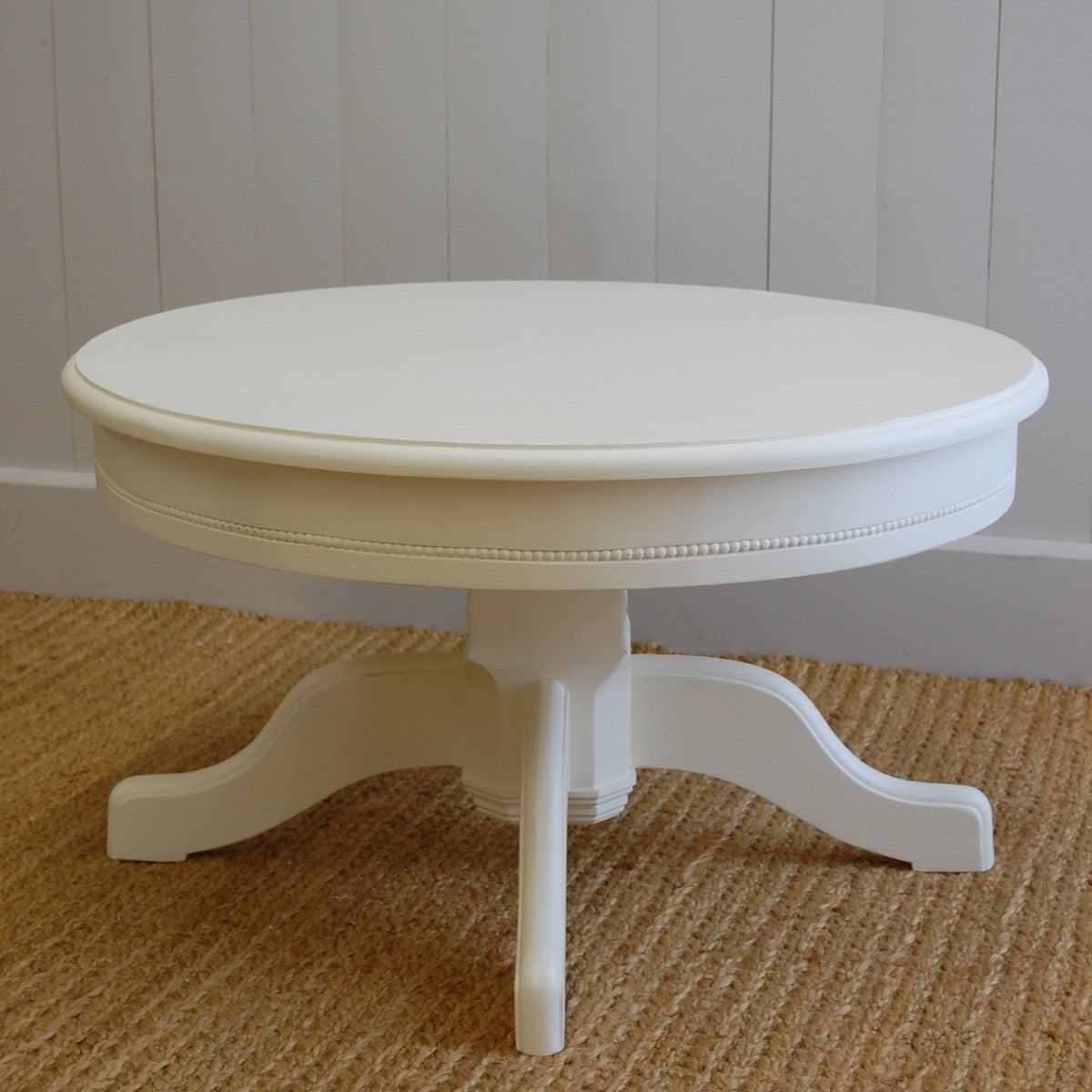 Off-white round wooden coffee table with beaded apron trim, a central pedestal and curved feet.