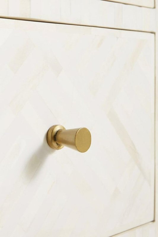 Macro close-up of a cylindrical solid brass knob on a white optical bone inlay dresser drawer, showcasing the premium hardware quality against the handcrafted bone inlay surface.