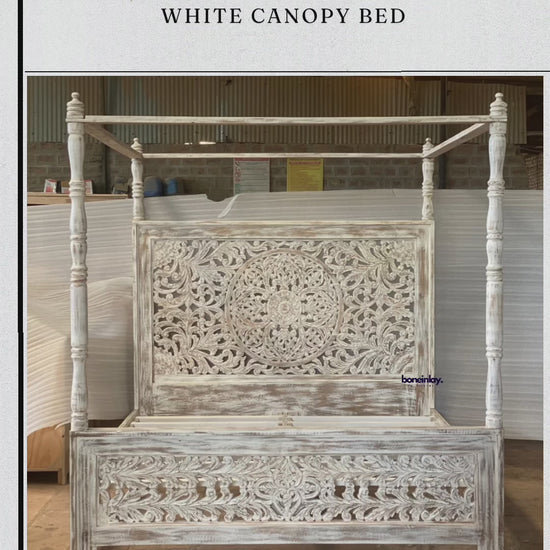 A factory video showcasing a hand-carved white canopy bed with a rustic farmhouse aesthetic. The design features an intricate floral mandala headboard and a coordinating footboard, all finished in a distressed "shabby chic" white paint. The video displays the solid wood construction, traditional Indian artistry, and four-poster frame, highlighting services like free worldwide delivery and customization.