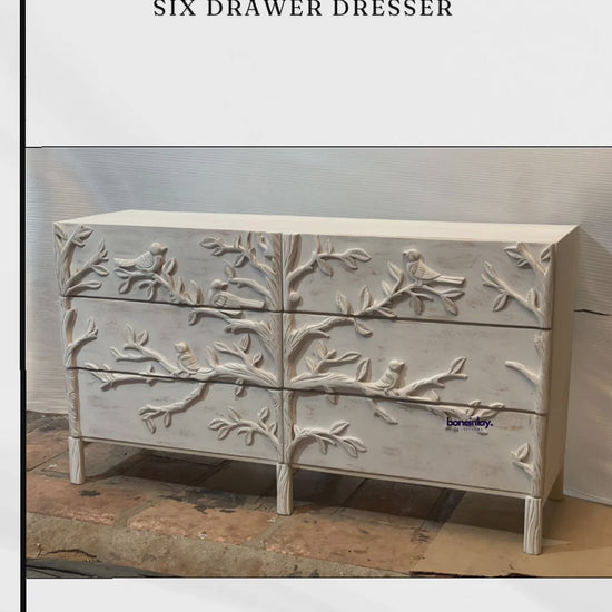 A wide six-drawer wooden dresser with a distressed whitewash finish. The entire front facade is covered in an intricate hand-carved relief design of birds on tree branches. The piece features a clean rectangular shape and sits on short, square-tapered legs, blending organic art with functional furniture design.