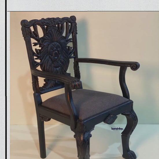 This video features a "Handcarved Menagerie Lion Dining Chair with Arm," showcasing a striking, hand-carved lion’s face as the centerpiece of the openwork backrest. The solid wood frame is finished in a deep, dark espresso tone that highlights the intricate artisanal textures, while the seat is upholstered in a smooth, charcoal gray fabric. Detailed close-ups emphasize the masterfully sculpted lion motif, elegant curved arms, and ornate cabriole legs, blending regal artistry with functional furniture design