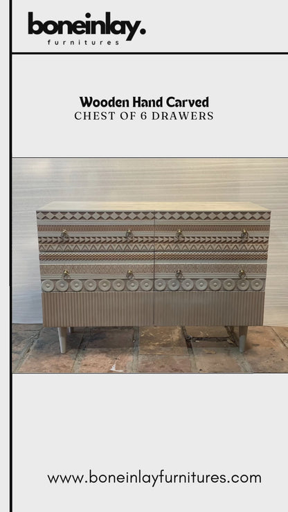 A wide, six-drawer wooden chest with intricate hand-carved geometric patterns on the drawer fronts. The light wood finish is accented by circular gold ring pulls and sits on four tapered white legs. The design features alternating rows of diamond, zigzag, and floral-inspired carvings