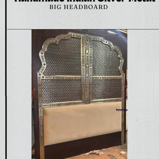 The "Handmade Indian Silver Metal Big Headboard" by boneinlay furnitures features an imposing, arched structure intricately decorated with traditional geometric openwork metal overlays. This elaborate silver pattern stands in contrast to the rich, dark wood frame, while the lower section is anchored by a smooth, cream-colored upholstered panel. The video showcases detailed close-ups of the fine metal embossing, demonstrating the craftsmanship from multiple angles.