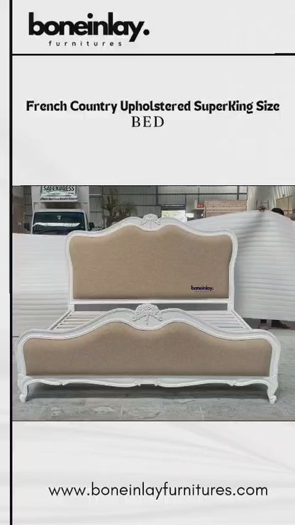 A French Country Upholstered Superking Size Bed from boneinlay furnitures featured in a showroom setting, highlighting its classic, ornate white wooden frame and textured beige upholstered headboard and footboard. The video pans around the bed to show intricate hand-carved floral accents on the crest, a slatted base structure, curved legs, and fine craftsmanship from multiple angles.