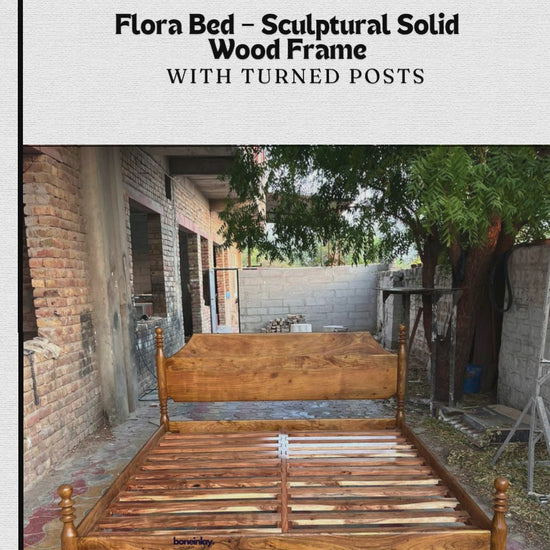 A promotional video showcases the Flora Bed, a wooden bed frame featuring a sleek, sculptural solid wood design with four elegant, turned posts at each corner. The video displays the empty wooden frame in an outdoor brick setting before transitioning to a fully furnished, styled bedroom, highlighting the bed's rustic yet refined aesthetic.